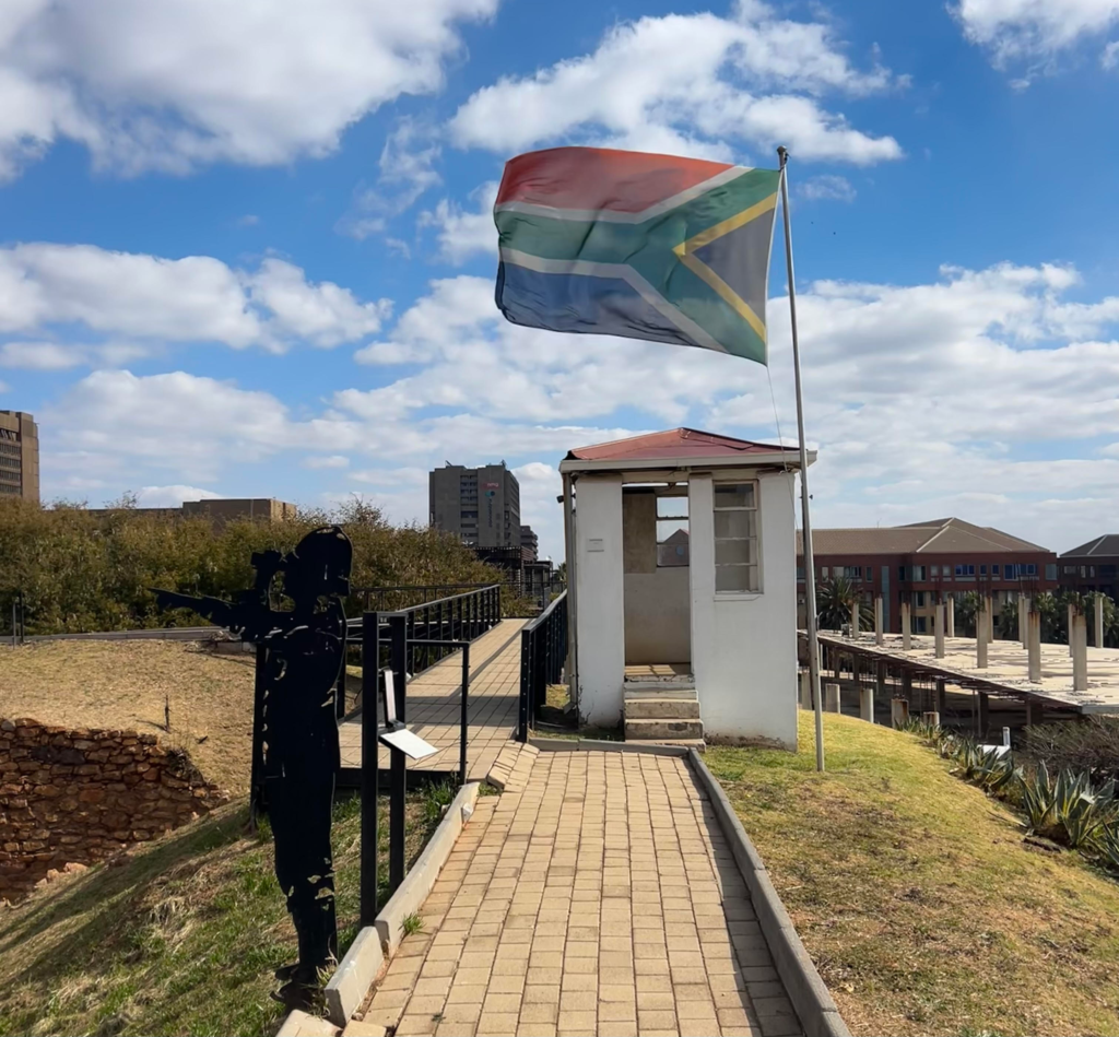 A 2D figure of a warden looking through binoculars with the South African flag in the back ground at Constitution Hill